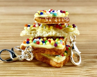 May include: A miniature stack of toast slices, each topped with yellow icing and colourful sprinkles. The toast has a golden-brown crust. A silver-coloured keychain clip is attached to the bottom slice.