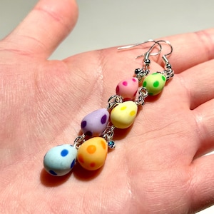 Peut inclure: Boucle d'oreille pendante avec six perles en forme d'œuf aux couleurs pastel, chacune avec des pois. Les couleurs sont le bleu, l'orange, le jaune, le violet, le rose et le vert. La boucle d'oreille est assemblée avec des chaînes et des crochets en métal argenté.