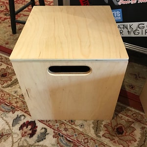 Puede incluir: Una caja de almacenamiento de madera clara con un asa en la parte delantera. La caja est&aacute; sentada sobre una alfombra estampada.