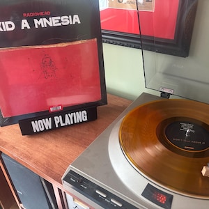 Puede incluir: Un tocadiscos con tapa transparente reproduce un disco de vinilo naranja translúcido. Una portada de álbum negra con el texto "RADIOHEAD KID A MNESIA" se encuentra sobre una superficie de madera, con un letrero "NOW PLAYING" delante.
