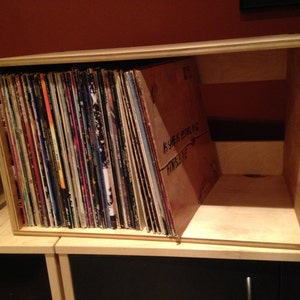May include: A wooden box with a shelf inside, filled with vinyl records. The box is made of light wood and has a natural finish. The records are stacked vertically and are visible through the open front of the box.
