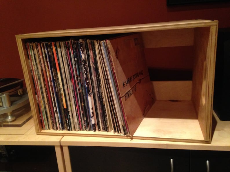 May include: A wooden box with a shelf inside, filled with vinyl records. The box is made of light wood and has a natural finish. The records are stacked vertically and are visible through the open front of the box.
