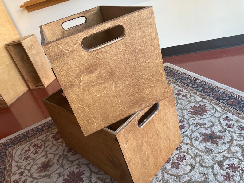 May include: Two wooden storage boxes with handles. The boxes are stacked on top of each other. The boxes are made of light brown wood and have a natural finish.