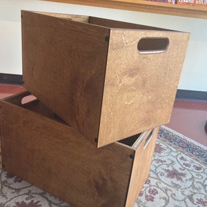 May include: Two wooden storage boxes stacked on top of each other. The boxes are made of brown plywood and have a natural wood finish. The top box has a cut-out handle on the front.