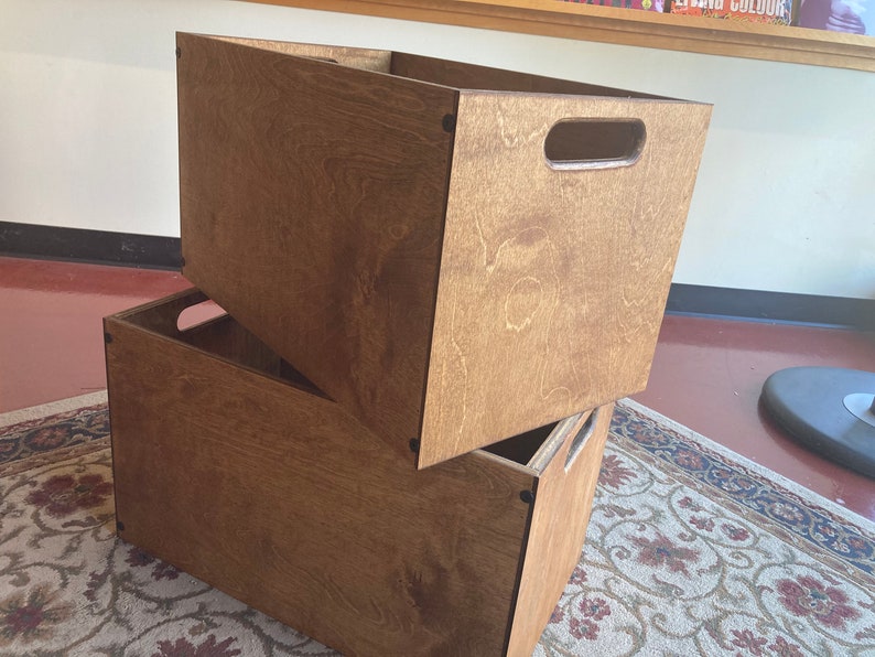 May include: Two wooden storage boxes stacked on top of each other. The boxes are made of brown plywood and have a natural wood finish. The top box has a cut-out handle on the front.