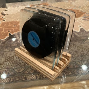 May include: Set of four glass coasters featuring a black vinyl record design with a blue center. The coasters are stacked on a wooden stand.