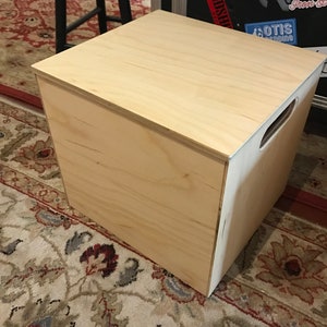 Puede incluir: Una caja de almacenamiento de madera clara con una tapa con bisagras y un asa en el lateral. La caja est&aacute; sobre una alfombra estampada.