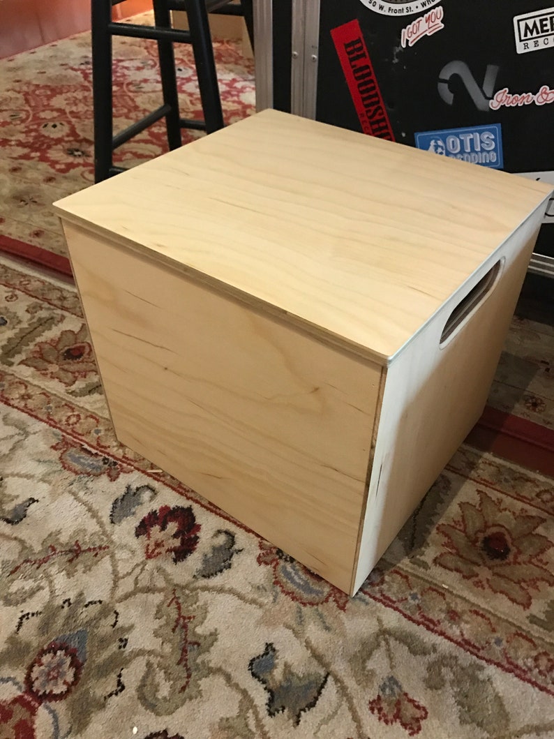 Puede incluir: Una caja de almacenamiento de madera clara con una tapa con bisagras y un asa en el lateral. La caja est&aacute; sobre una alfombra estampada.