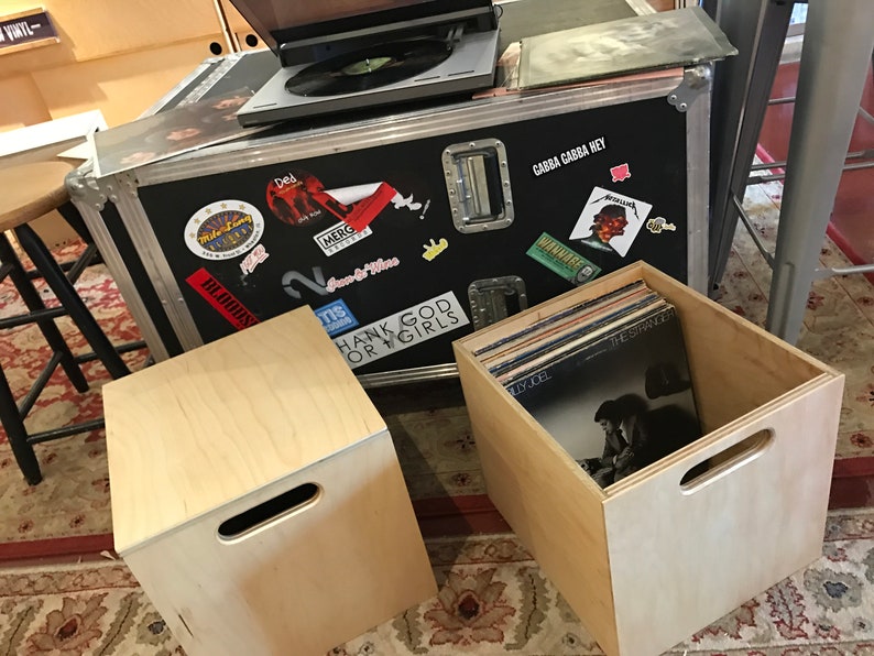Puede incluir: Dos cajas de madera con asas, una llena de discos de vinilo. La caja con los discos tiene el texto "Billy Joel" y "The Stranger" visible en las portadas de los discos. La otra caja est&aacute; vac&iacute;a. Las cajas est&aacute;n sobre una alfombra estampada delante de una caja de metal negra con pegatinas.