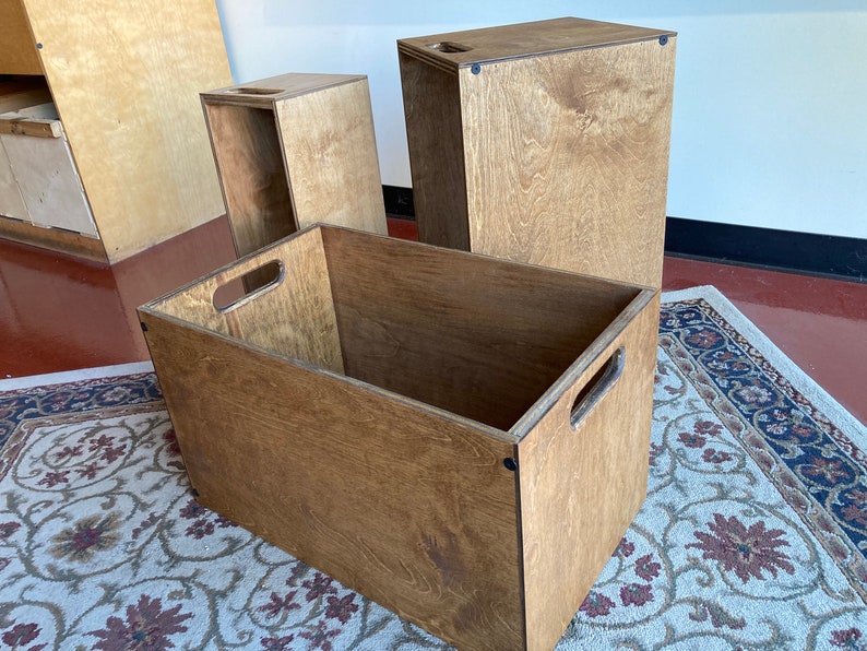 May include: Three wooden storage boxes with lids. The boxes are made of plywood and have a natural wood finish. The largest box is on the left, followed by a smaller box, and then the smallest box on the right.