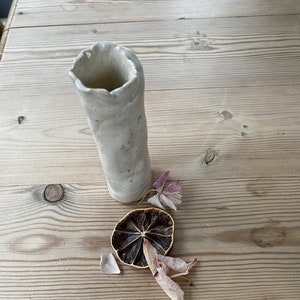 May include: A white ceramic vase with a rough, textured surface. The vase is sitting on a wooden table with dried flowers and a dried lemon slice.
