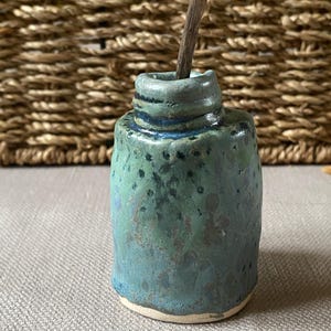 May include: A small, teal blue ceramic vase with a white rim and a speckled glaze. The vase is sitting on a woven surface.