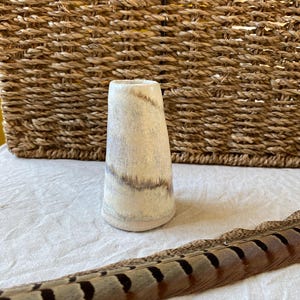 May include: A small, off-white ceramic vase with brown and grey streaks. The vase has a slightly flared shape and sits on a white surface. A brown and black feather lies in the foreground, with a woven basket in the background.