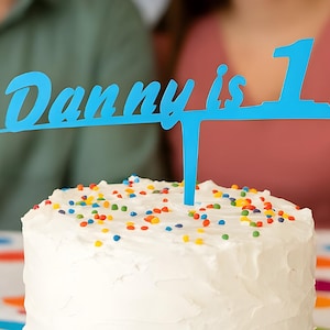 Puede incluir: Un pastel redondo glaseado blanco con base de chocolate y borde de glaseado azul. El pastel está decorado con chispas de colores y un adorno azul que dice "Danny is 1".