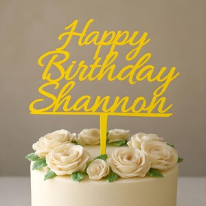 Puede incluir: Un adorno para pastel amarillo con las palabras "Happy Birthday Shannon" en cursiva. El pastel está decorado con rosas blancas y hojas verdes. El pastel tiene un glaseado blanco.