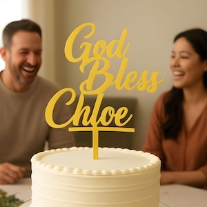 Puede incluir: Un pastel blanco glaseado con un adorno amarillo que dice "God Bless Chloe". El pastel está sobre un soporte blanco y dorado. La imagen también incluye el texto "Had it Made Design Studio".