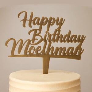 Puede incluir: Un adorno dorado para pastel con las palabras "Happy Birthday Moelhman" en una fuente cursiva. El adorno está insertado en un pastel glaseado blanco con un diseño acanalado. El fondo es de color beige claro sólido.