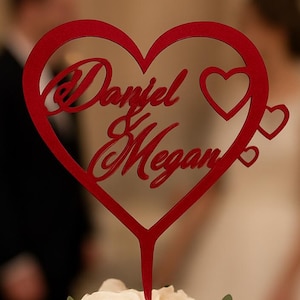 Puede incluir: Un adorno para pastel rojo en forma de corazón con los nombres "Daniel & Megan" en escritura cursiva. El adorno está sobre un pastel blanco de varios niveles decorado con rosas blancas y vegetación. El fondo está borroso, mostrando una escena de boda.