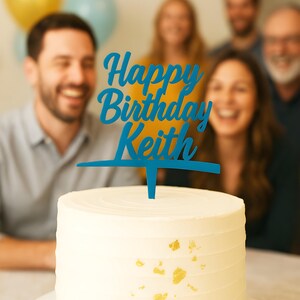 Puede incluir: Un pastel blanco glaseado con detalles de hojas doradas, coronado con un adorno azul que dice "Happy Birthday Keith". Varias personas están borrosas en el fondo, con globos en la esquina superior izquierda.