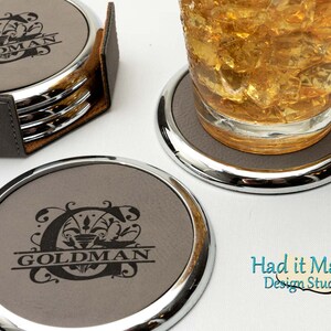Puede incluir: Conjunto de cuatro posavasos de cuero gris con borde de metal plateado. Cada posavasos presenta un monograma personalizado con el nombre "GOLDMAN" en una fuente decorativa. Los posavasos están apilados en un soporte.