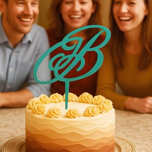 Pode incluir: Um bolo redondo com um glacê gradiente marrom, bege e amarelo, coberto com redemoinhos de creme de manteiga amarelos e um topo de bolo turquesa com a letra "B". O bolo está em um prato decorativo.