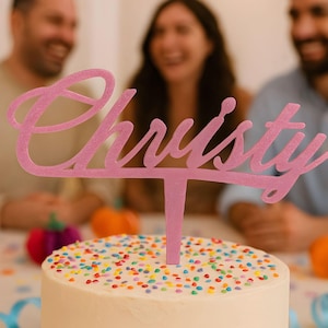 Puede incluir: Un adorno para pastel rosa brillante con el nombre "Christy" en escritura cursiva. El adorno está colocado sobre un pastel glaseado blanco cubierto de chispas de colores. Personas borrosas en el fondo sugieren una celebración.