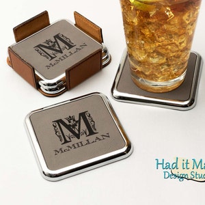 Puede incluir: Conjunto de cuatro posavasos cuadrados grises con un borde plateado. Cada posavasos presenta un monograma personalizado con la inicial "M" y el nombre "McMillan" grabado en negro. Los posavasos están apilados en un soporte de cuero.