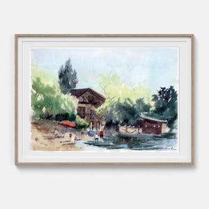 Könnte beinhalten: Ein gerahmtes Aquarellbild, das eine Seeuferlandschaft mit einem Holzhaus, üppigen grünen Bäumen und einem Sandstrand zeigt. Mehrere kleine Figuren sind in der Nähe des Wassers zu sehen, zusammen mit einem roten Sonnenschirm am Ufer.