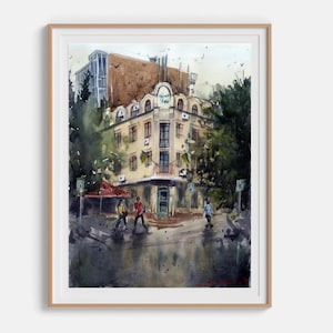 Könnte beinhalten: Ein Aquarellgemälde eines mehrstöckigen Gebäudes mit abgerundeter Eckfassade, das Bogenfenster und einen zentralen Eingang aufweist. Die Szene umfasst dunkelgrüne Bäume, ein rotes Vordach und auf der Straße gehende Figuren, gerahmt in hellem Holz.