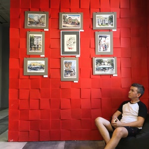 May include: A young person sits in a beanbag chair in front of a red wall with framed watercolor paintings of cityscapes. The paintings are arranged in a grid pattern.