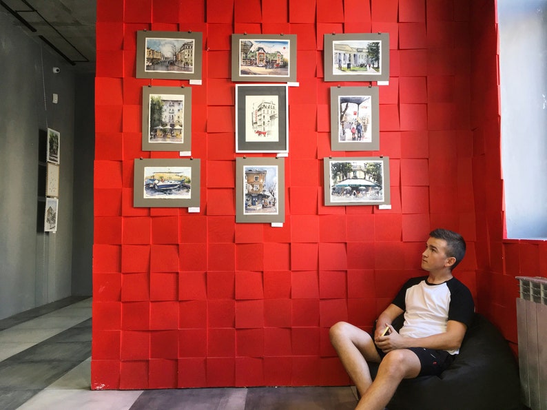 May include: A young person sits in a beanbag chair in front of a red wall with framed watercolor paintings of cityscapes. The paintings are arranged in a grid pattern.