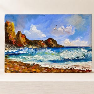 May include: An oil painting on canvas depicting a coastal scene with a rocky shoreline, a pebble beach in the foreground, and crashing blue waves. A single white bird flies in a bright blue sky filled with white clouds above the dark cliffs.
