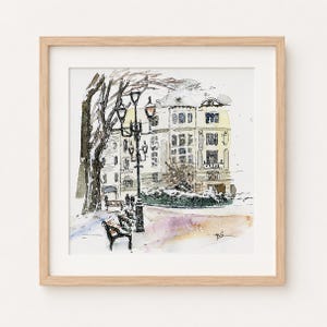 May include: A framed watercolor painting depicting a snowy city scene with a tall, multi-story building, a dark ornate street lamp, and a park bench in the foreground. The artwork features muted yellow and brown tones with black ink outlines.