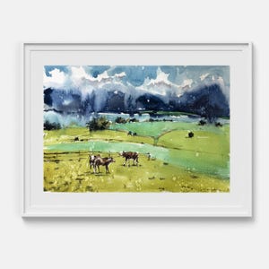 May include: A framed watercolor painting depicting a rural landscape with rolling green hills and two brown and white cows standing in a grassy pasture. The background features dark blue, stormy clouds hanging over distant mountains and fields.