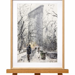 May include: Watercolor painting of a snowy city scene, framed in light wood. The artwork depicts a street with pedestrians, bare trees, and a tall building in the background. The painting is displayed on a wooden easel.