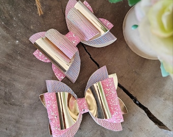May include: Two pink and gold glitter hair bows with a layered design. The bows have a metallic gold center and are decorated with iridescent glitter.