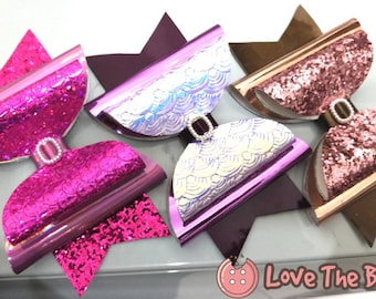 May include: Three hair bows with a star-shaped base. The bows are made of iridescent material with a glitter finish. The bows are in pink, purple, and rose gold.