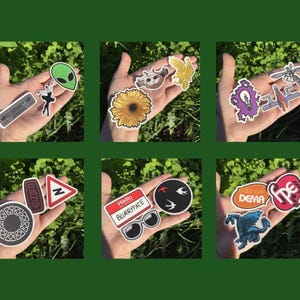 May include: A collection of colorful stickers featuring various designs, including an alien, a sunflower, and text that says "Hello BLURRYFACE". The stickers are held in a hand against a green background.