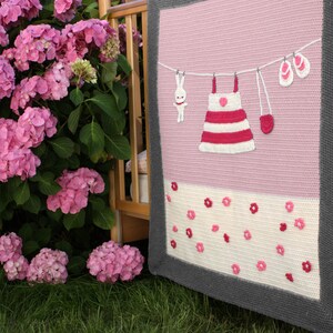 Puede incluir: Una manta tejida a mano en rosa y blanco con borde gris. Presenta un tendedero con un vestido, un conejito, mitones y sandalias. La secci&oacute;n inferior tiene tela blanca con detalles de flores rosas. Hecho a mano.