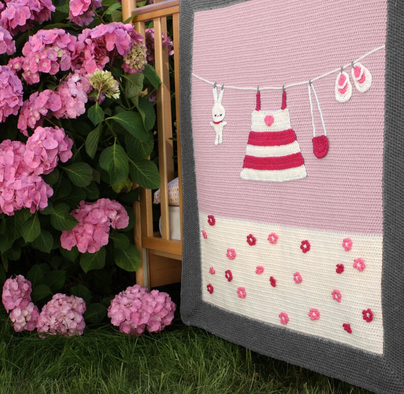 Puede incluir: Una manta tejida a mano en rosa y blanco con borde gris. Presenta un tendedero con un vestido, un conejito, mitones y sandalias. La secci&oacute;n inferior tiene tela blanca con detalles de flores rosas. Hecho a mano.