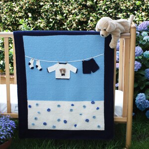 Puede incluir: Una manta de beb&eacute; hecha a mano en tonos azules y blancos, con un borde azul oscuro. La manta presenta aplicaciones de ropa de ganchillo, incluyendo un su&eacute;ter con un oso de peluche, pantalones cortos y calcetines, colgados en un tendedero. Un cachorro de peluche descansa en la cuna.