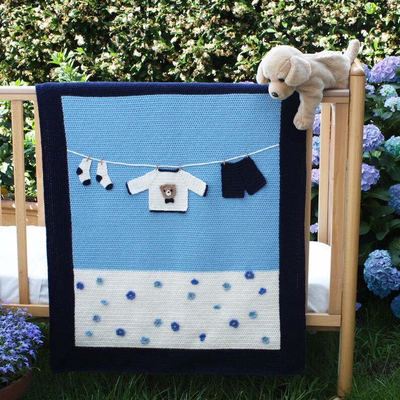 Puede incluir: Una manta de beb&eacute; hecha a mano en tonos azules y blancos, con un borde azul oscuro. La manta presenta aplicaciones de ropa de ganchillo, incluyendo un su&eacute;ter con un oso de peluche, pantalones cortos y calcetines, colgados en un tendedero. Un cachorro de peluche descansa en la cuna.