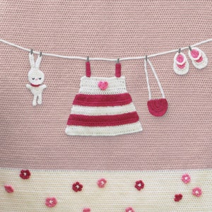 Puede incluir: Un dise&ntilde;o de ganchillo con un conejito blanco, un vestido a rayas con un coraz&oacute;n, un peque&ntilde;o bolso y zapatos de beb&eacute; colgados de un tendedero. El dise&ntilde;o est&aacute; sobre un fondo rosa con detalles florales en la parte inferior.