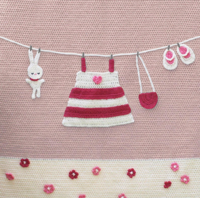 Puede incluir: Un dise&ntilde;o de ganchillo con un conejito blanco, un vestido a rayas con un coraz&oacute;n, un peque&ntilde;o bolso y zapatos de beb&eacute; colgados de un tendedero. El dise&ntilde;o est&aacute; sobre un fondo rosa con detalles florales en la parte inferior.