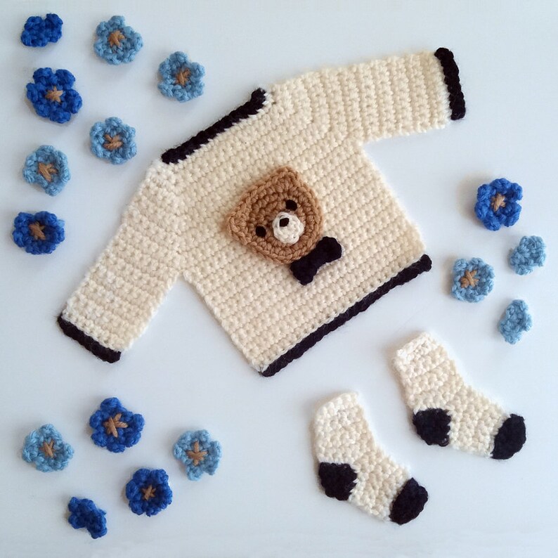 Puede incluir: Un conjunto de beb&eacute; de ganchillo color crema, que incluye un su&eacute;ter con un aplique de oso marr&oacute;n y una pajarita negra, y calcetines a juego. Flores azules y crema de ganchillo est&aacute;n dispersas alrededor.