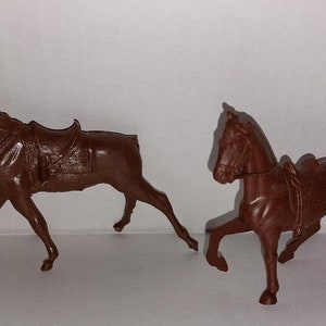 May include: Two brown plastic toy horses with saddles. The horses are in a walking pose.