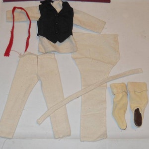 May include: A set of doll clothes for a historical figure, including a white shirt, black vest, white pants, a red sash, and a pair of white boots.