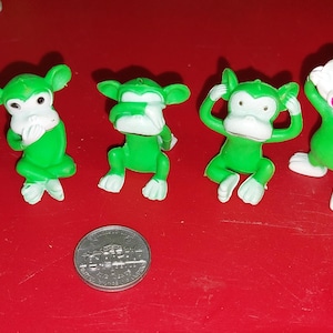 May include: Five small green monkey figurines with white faces, hands, and feet, each in a different pose. One monkey lies down, one covers its mouth, one covers its eyes, one covers its ears, and one stands with arms raised. A US nickel is shown for scale.