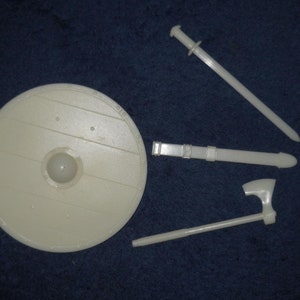 May include: A white plastic shield, sword, and axe. The shield is round and has a raised center. The sword has a simple handle and blade. The axe has a wooden handle and a metal blade.