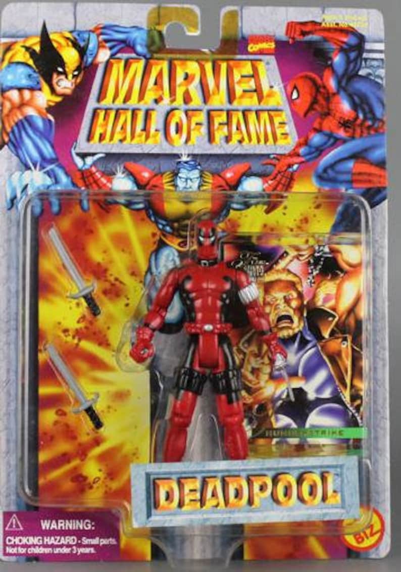 May include: A red and black action figure of Deadpool from the Marvel Hall of Fame series. The figure is holding two swords and is standing on a base with the word "DEADPOOL" printed on it.